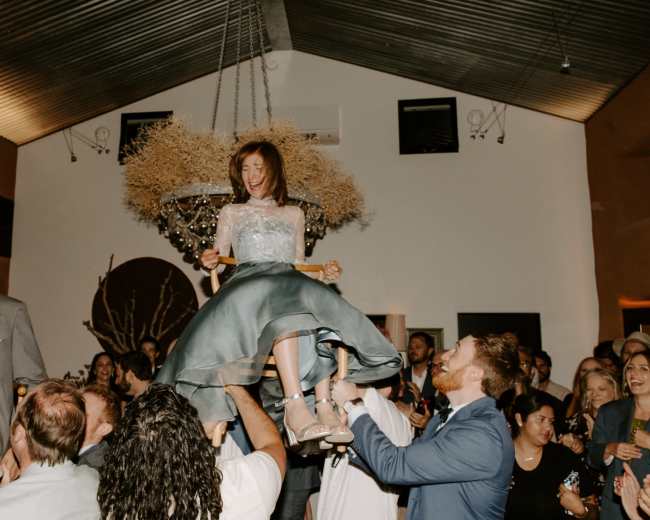 A woman in a formal dress is being lifted into the air by several people during a celebration inside a room with high ceilings and decorative lighting.