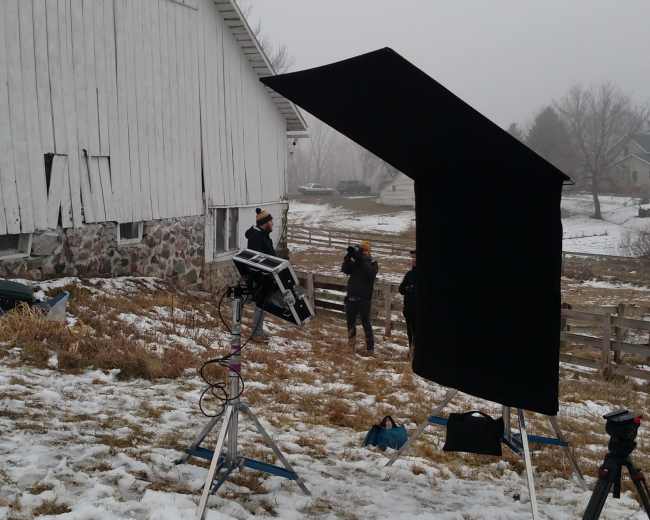 A film crew is setting up equipment outside a barn on a snowy field.