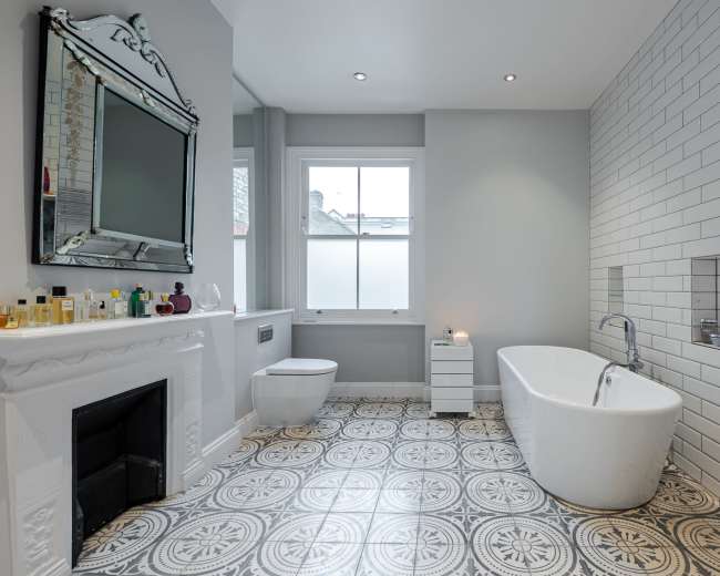 A modern bathroom features a freestanding bathtub, a sleek toilet, and a large mirror above a decorative fireplace, with patterned tiles on the floor and a window allowing natural light.