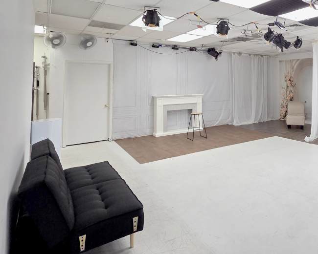 The image shows a well-lit photography studio with a white backdrop, a black couch, a stool, and various lighting equipment set up against a plain wall.