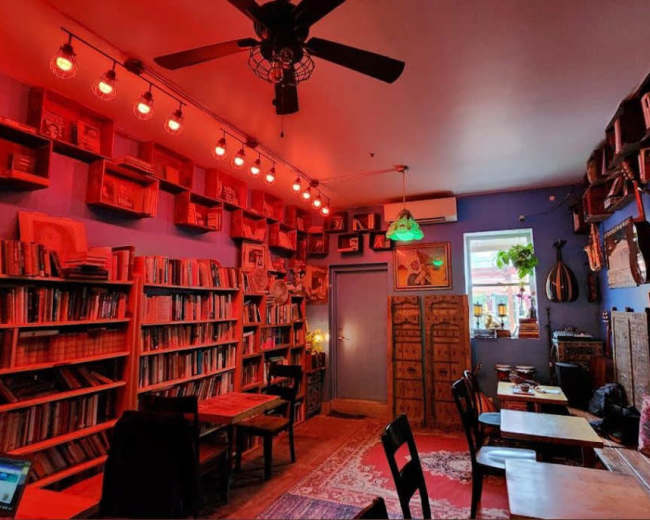 The image depicts a cozy bookshop with red accent lighting, filled with bookshelves lining the walls and wooden tables scattered throughout the space.
