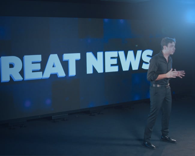 A person stands on a stage in front of a large screen displaying the text "GREAT NEWS."