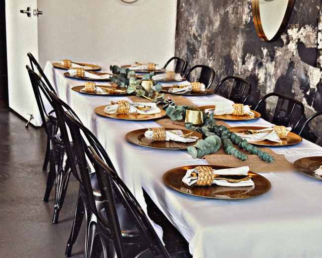 A long dining table is set with place settings featuring plates, napkins, and decorative elements, surrounded by black chairs in a modern interior space.