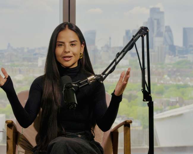 A woman with long black hair is seated in a chair with a microphone in front of her, gesturing while overlooking a city skyline through a large window.