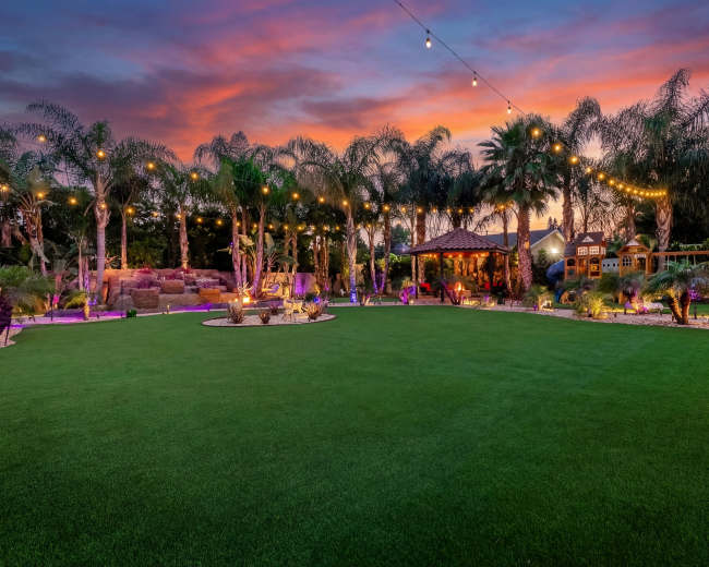 A lush green backyard is illuminated by string lights, featuring palm trees, a gazebo, and a play structure against a vibrant sunset.