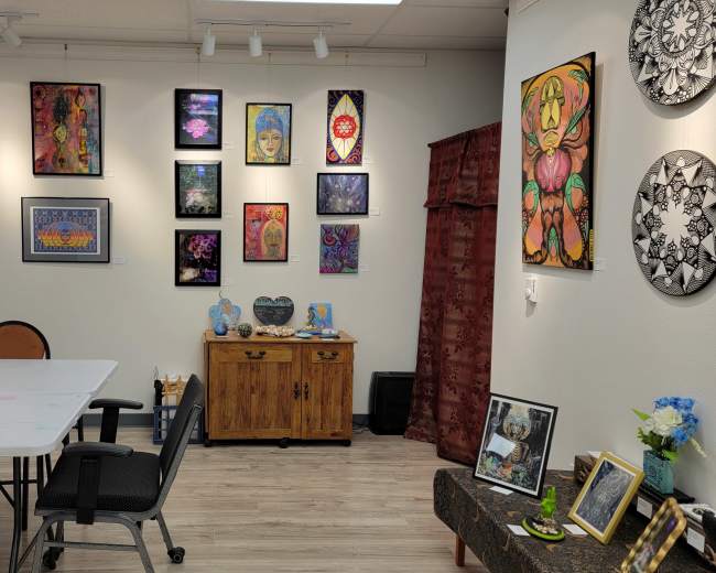 The image shows an art studio with a large table in the center, surrounded by chairs, and walls decorated with colorful paintings and artwork.