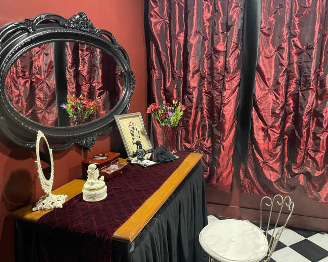 The image features a vintage vanity table with a mirror, surrounded by red velvet curtains and a decorative chair on a black and white checkered floor.