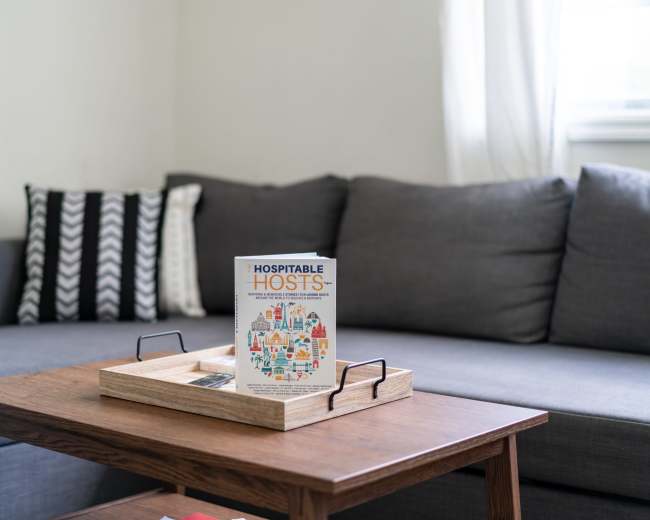 A coffee table with a wooden tray displays a book titled "Hospitable Hosts" in front of a gray sofa with patterned cushions.