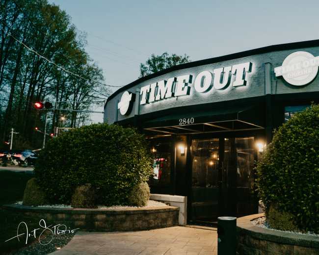The image shows the exterior of a restaurant named "TIME OUT" with a well-lit sign and neatly manicured bushes in front.