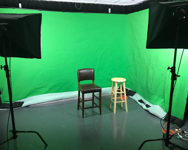 A studio setup with a green screen backdrop, featuring a brown wooden chair and a yellow stool positioned in front of it.