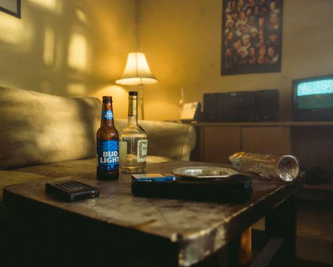 A low-lit living room scene with a coffee table containing two beer bottles, a liquor bottle, a cigarette ashtray, and a remote, while a lamp and a small television are in the background.