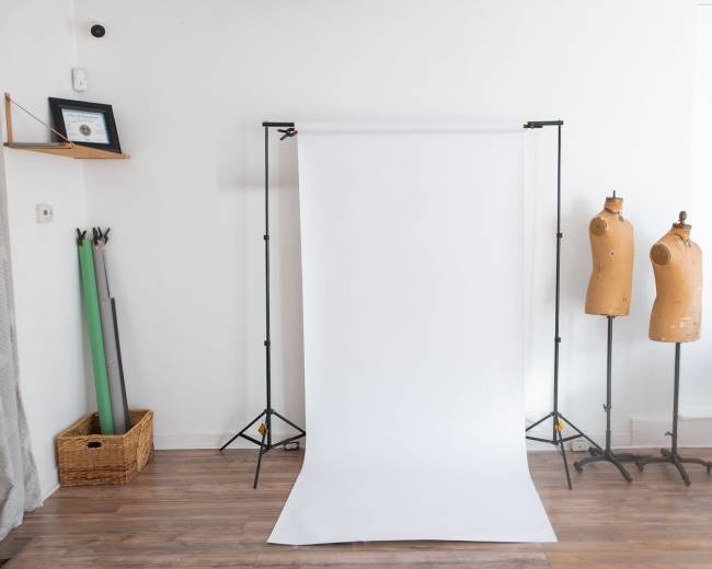 The image shows a photography setup with a white backdrop on stands, two dress forms on the right, and some green props in a basket on the left.
