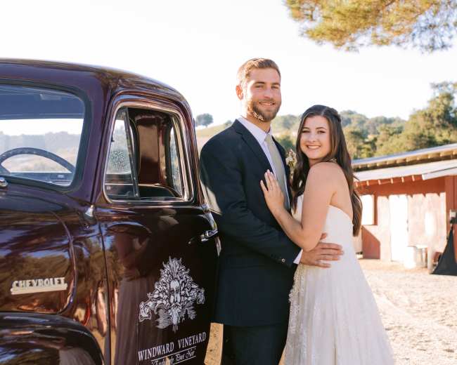 A couple poses happily next to a vintage Chevrolet truck at a vineyard.