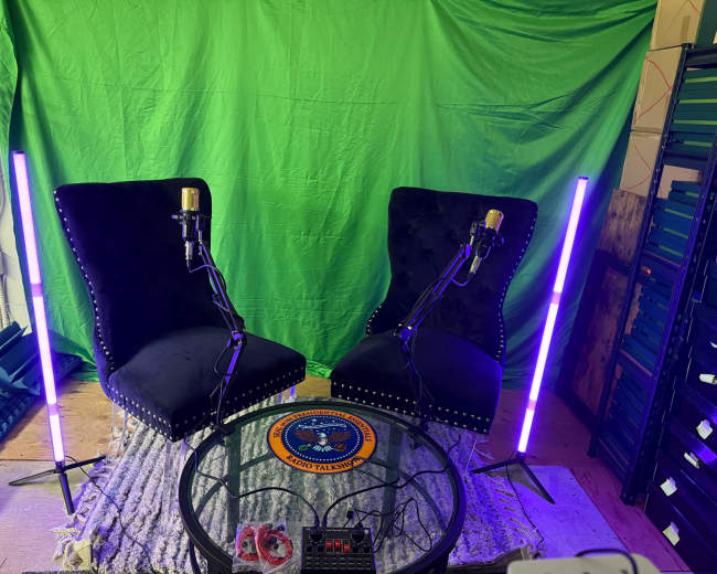 The scene features two black armchairs with microphones positioned on them, set against a green backdrop, with two illuminated purple light stands on either side of a round glass table.