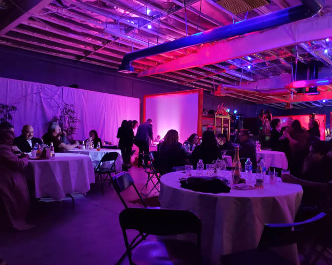 A dimly lit venue features several tables with guests dining, surrounded by colorful lights and a backdrop of plants.