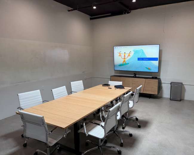 A modern conference room features a large wooden table surrounded by white chairs, with a wall-mounted screen displaying a colorful presentation.