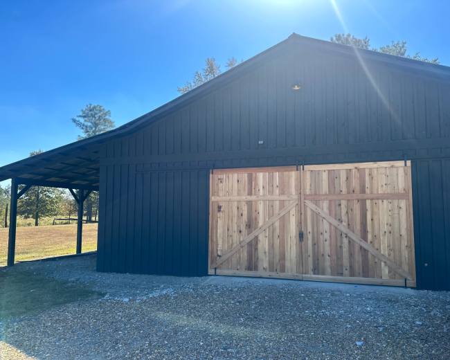 A dark wooden barn with a large wooden double door and a gravel pathway in front of it.