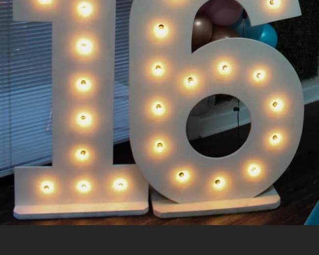 Large illuminated numbers "16" made of white material with bulbs, set against a backdrop of balloons and a wall.