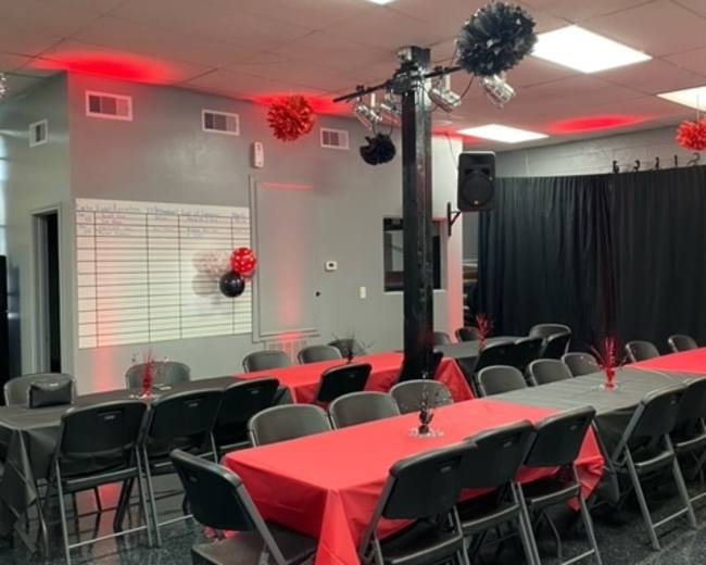 A room set up for an event, featuring several long tables covered with red tablecloths and black decorations hanging from the ceiling.