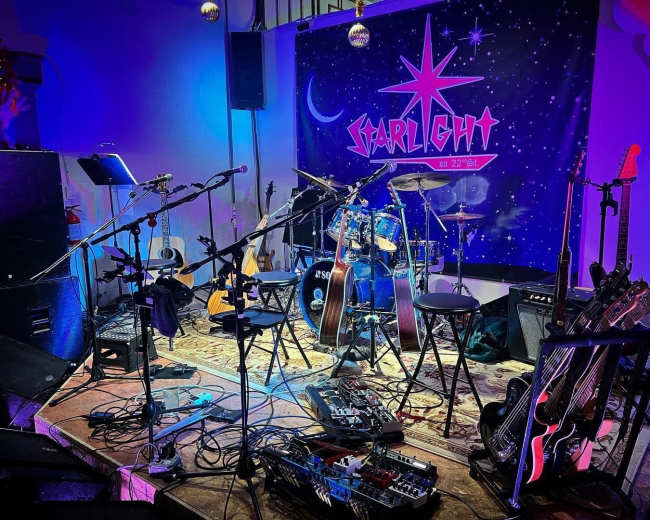 A stage setup featuring various musical instruments, including guitars and a drum kit, with a backdrop displaying the word "StarLight" and colorful lighting.