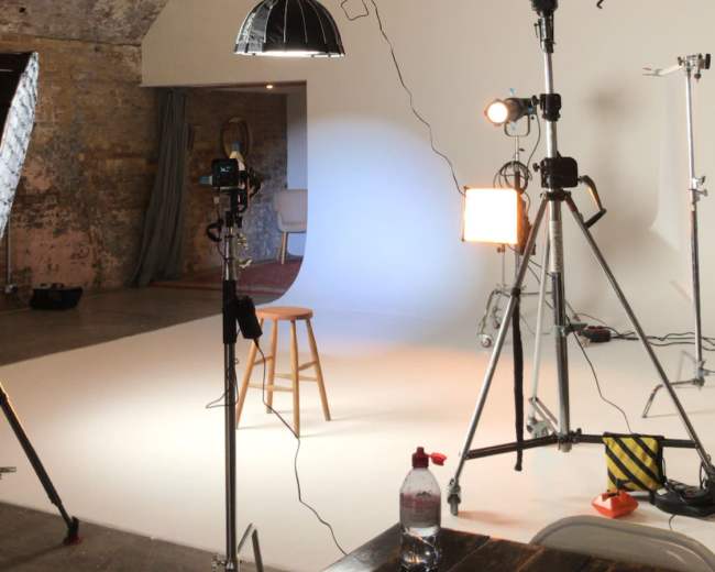 The image shows a photographic studio with a wooden stool, various lighting equipment, and a backdrop set up in an arched brick room.