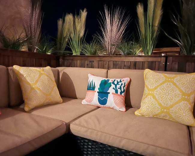 A couch with beige cushions and colorful throw pillows is situated in a dimly lit outdoor space with decorative plants in the background.