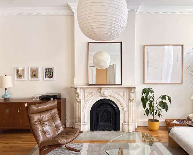 The image shows a living room featuring a brown leather chair, a modern lamp, a marble fireplace, a large round paper lantern, and decorative artwork on the walls.