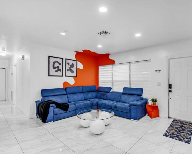 The image shows a modern living room featuring a blue sectional sofa, a round glass coffee table, a white door, and an accent wall painted in orange.