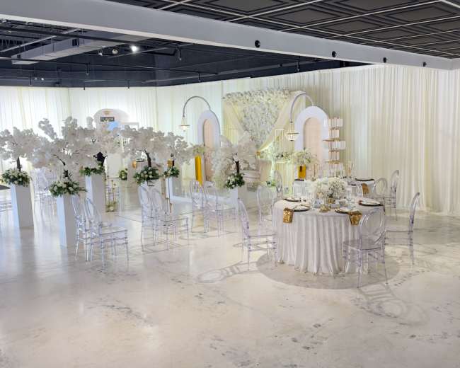 The image shows a decorated event space featuring elegant tables with white floral arrangements, transparent chairs, and a backdrop of draped white fabric.