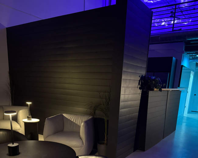 The image shows a modern interior space featuring a black wall, a round table with chairs, and soft lighting.