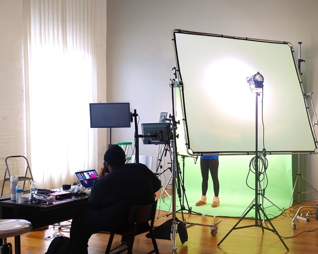 A person sits at a table in a studio, working on a computer in front of a green screen setup with lighting equipment behind them.