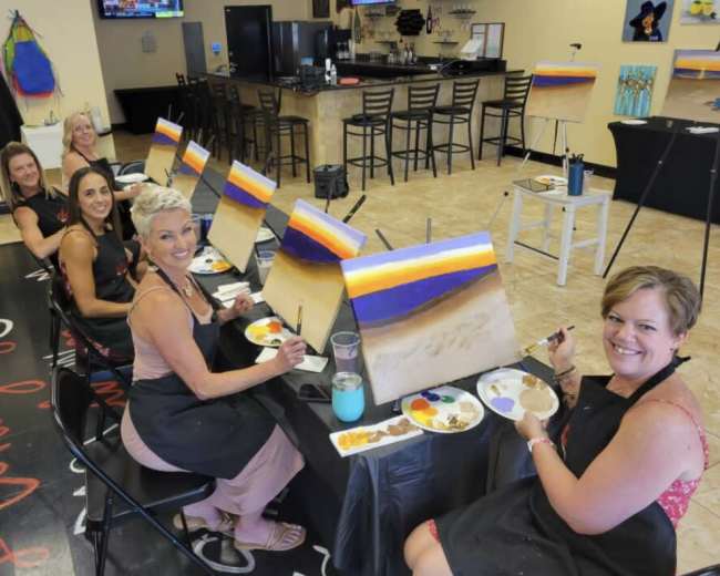 A group of six women is sitting at a table in a studio, each painting a canvas with a landscape, while various art supplies are arranged in front of them.