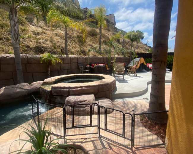 The image shows a backyard pool area with a hot tub, palm trees, and a fenced-off section on a sunny day.