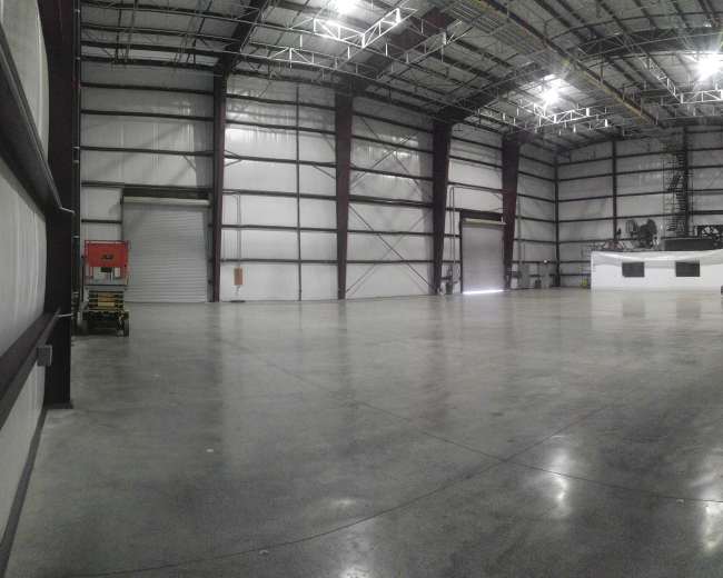 The image shows a large, empty industrial warehouse with high ceilings and a smooth concrete floor.