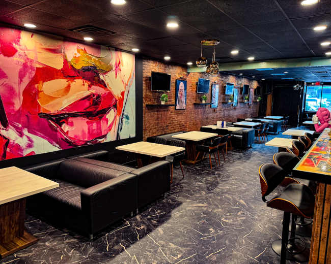 The image shows a modern café interior with a vibrant mural on one wall, featuring a close-up of a woman's face, and several tables and chairs arranged throughout the space.