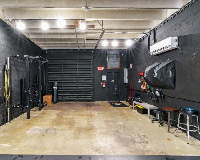 The image shows an empty gym space with black walls, various weight plates on the left, and fitness equipment along the right side.