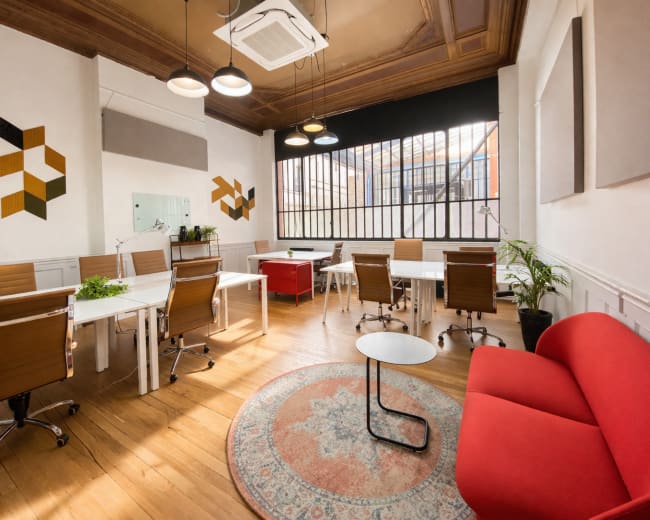 The image shows a modern workspace with several desks, ergonomic chairs, a red seating area, and large windows allowing natural light to fill the room.