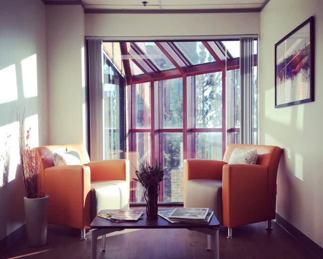 A brightly lit corner of an office featuring two orange chairs, a low coffee table with magazines, and a large window providing a view of the outdoors.
