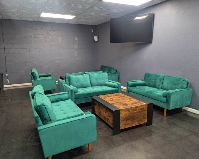 The image shows a waiting area with several green upholstered couches and a wooden coffee table arranged in an open space with a television mounted on the wall.