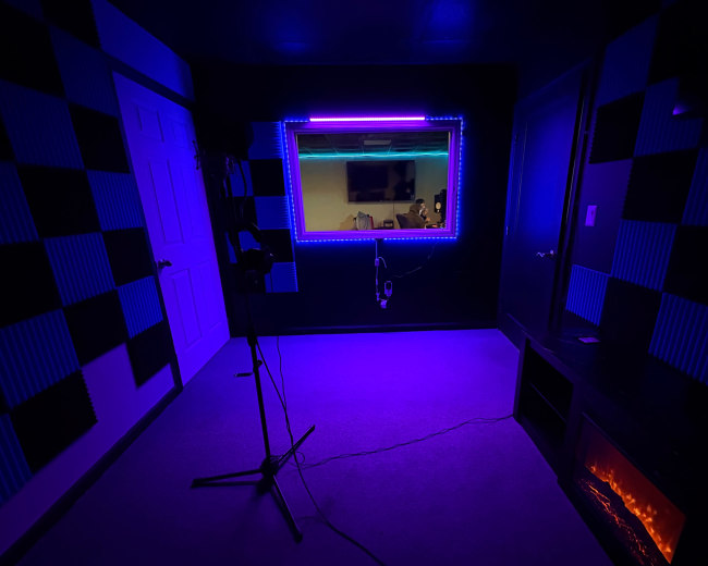 A dimly lit room features black and blue checkered walls, a microphone stand, and a brightly illuminated window with blue and purple LED lights.