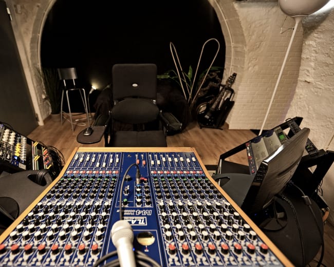 The image shows a sound mixing studio with a large mixing console at the center, flanked by two monitors, and a microphone positioned in front of a black chair.