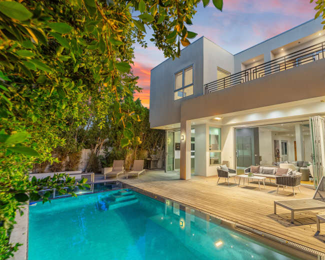 A modern house with large windows overlooks a swimming pool surrounded by wooden decking and lounge chairs, set against a colorful sunset sky.