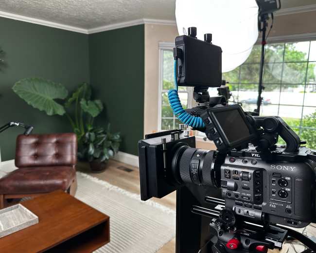 A camera is positioned on a tripod in a well-lit living room featuring a green accent wall, a brown leather chair, and several indoor plants.