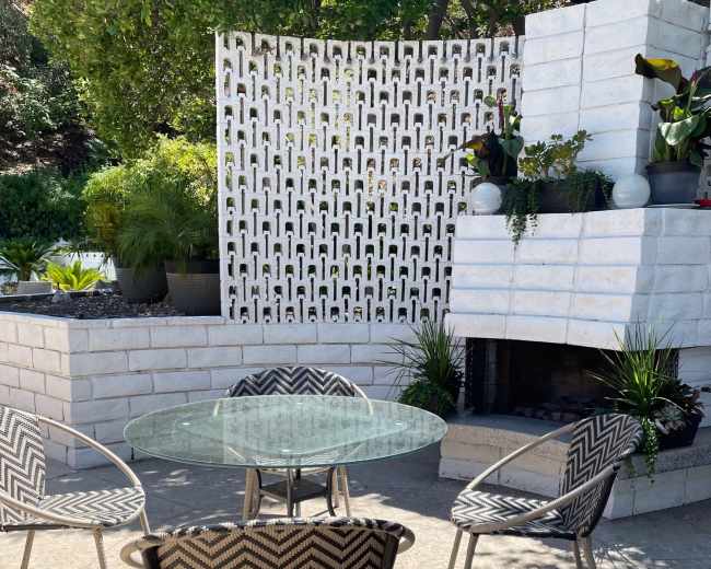 A circular glass table is surrounded by four patterned chairs on a patio with a white decorative wall and a fireplace.