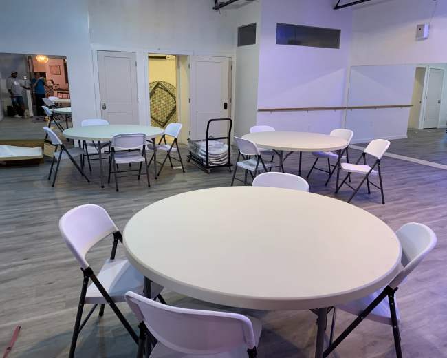 A spacious room with multiple round tables and folding chairs arranged for an event, with mirrors and a doorway visible in the background.