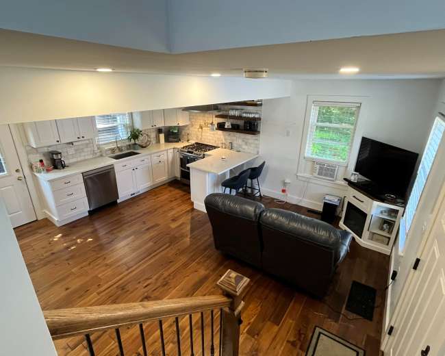 The image shows a spacious kitchen and living area with wooden floors, featuring a sofa, a TV stand, and a counter with bar stools.