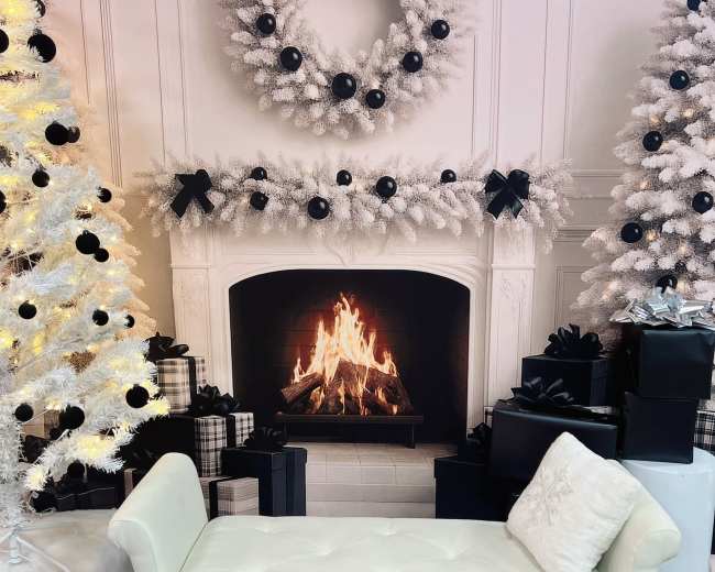 A white upholstered bench is positioned in front of a lit fireplace, surrounded by decorated Christmas trees and wreaths.