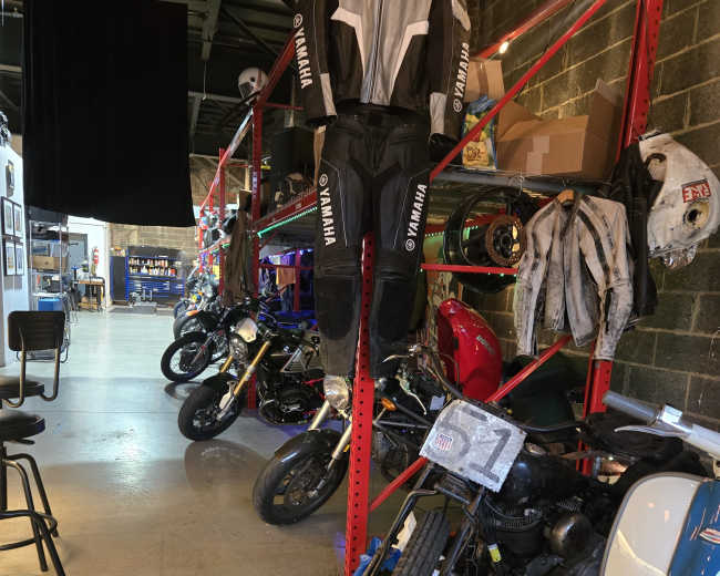 The image shows a garage filled with various motorcycles and race gear, including a Yamaha racing suit hanging on a rack.