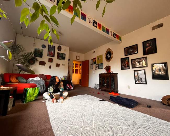 A person sits on the floor of a living room with a textured rug, surrounded by various artworks and decorations on the walls.