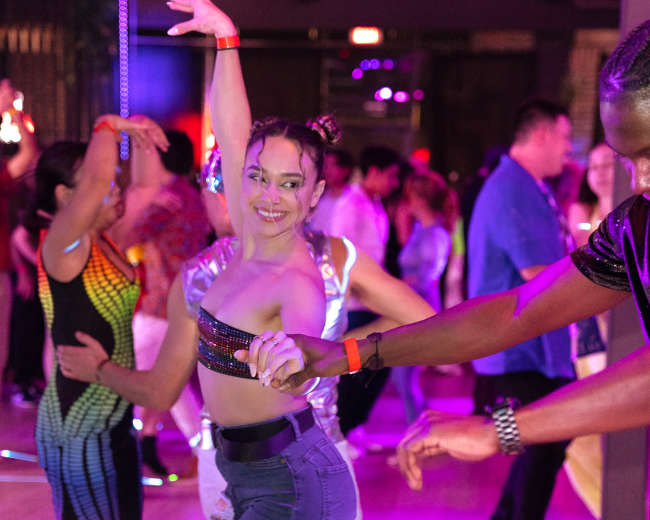 A group of people dances energetically in a dimly lit venue adorned with colorful lights.